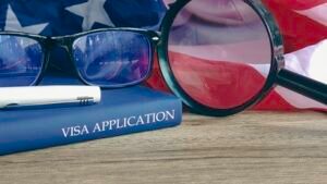 Magnifying glass and glasses on a book titled "VISA APPLICATION" with the American flag in the background.